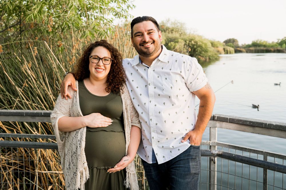 Maternity portrait of couple standing by water with reeds and desert scenery.