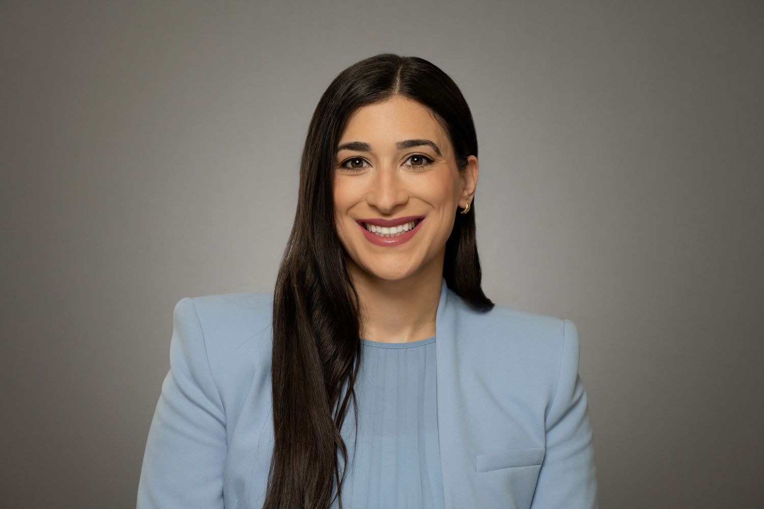 Smiling woman with long dark hair wearing a light blue blazer against a neutral gray background.
