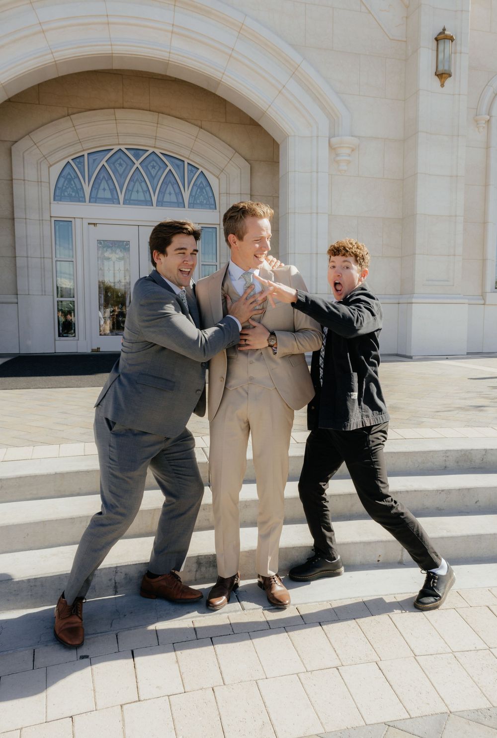 Three young men in suits playfully pose together on temple steps during wedding celebration.