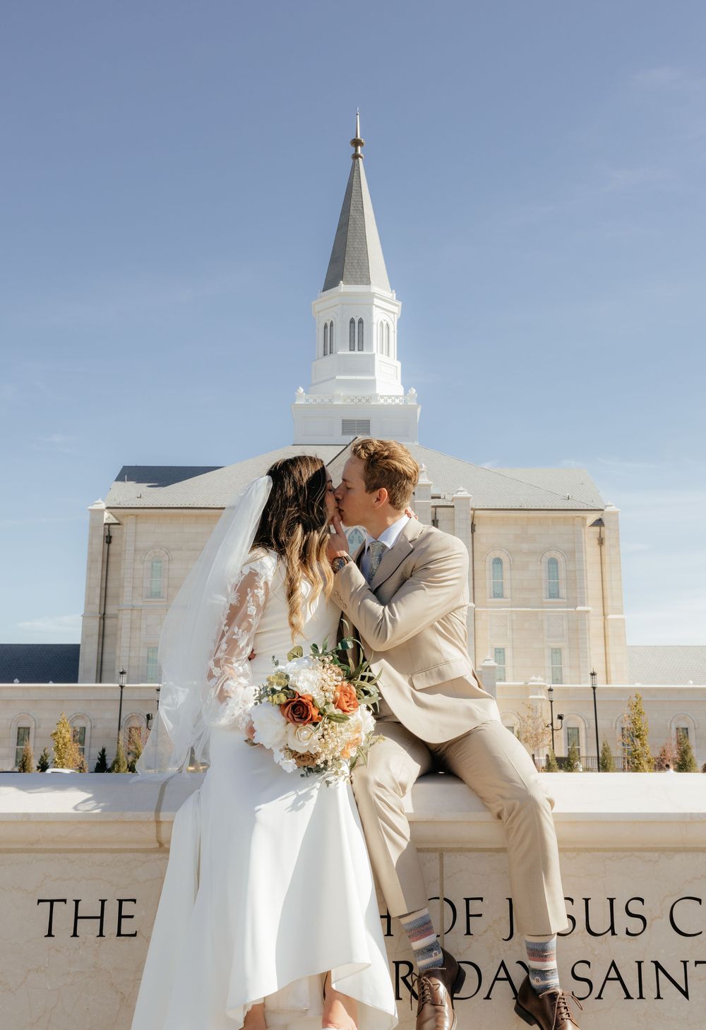 Bride and groom share a romantic kiss on temple grounds with white steeple against blue sky.