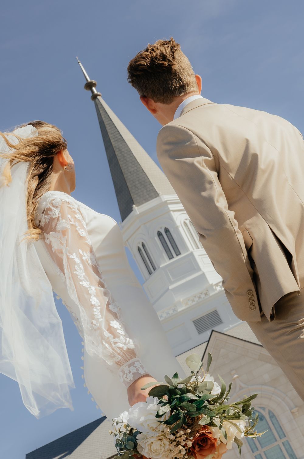 Bride and groom gaze up at LDS temple steeple, bride holding colorful bouquet on wedding day.