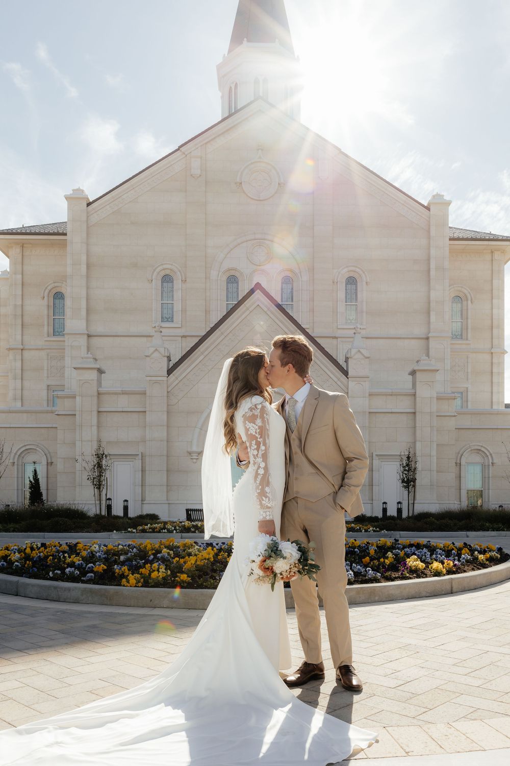 Bride and groom share a kiss in front of LDS temple with beautiful sun flare backdrop.