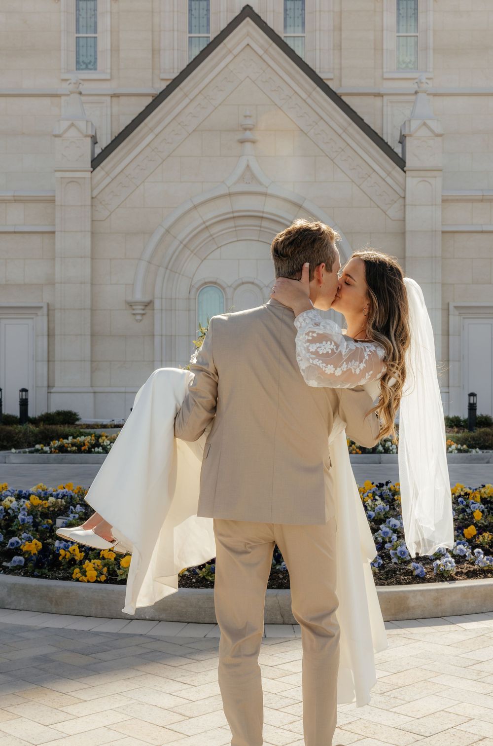 Groom carries bride in his arms sharing a kiss in front of blooming temple flower garden.