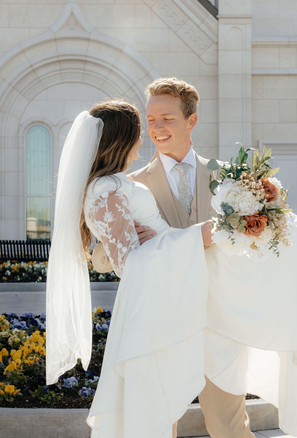 Smiling groom holds bride close outdoors near LDS temple flower garden on their wedding day.