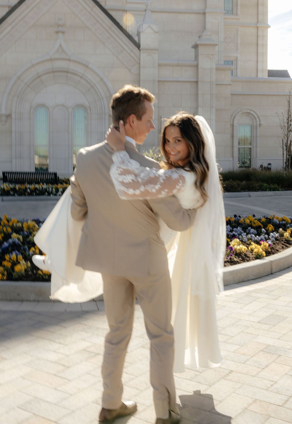 Bride and groom embracing outside a white LDS temple surrounded by colorful flower gardens on their wedding day.