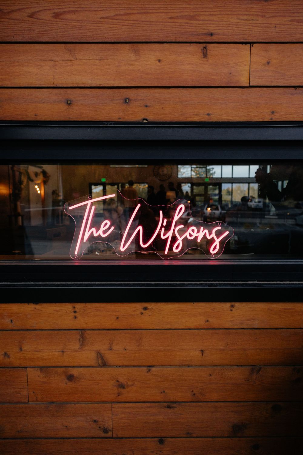 Pink neon sign reading The Wilsons mounted on a rustic wood-paneled wall at a wedding reception.