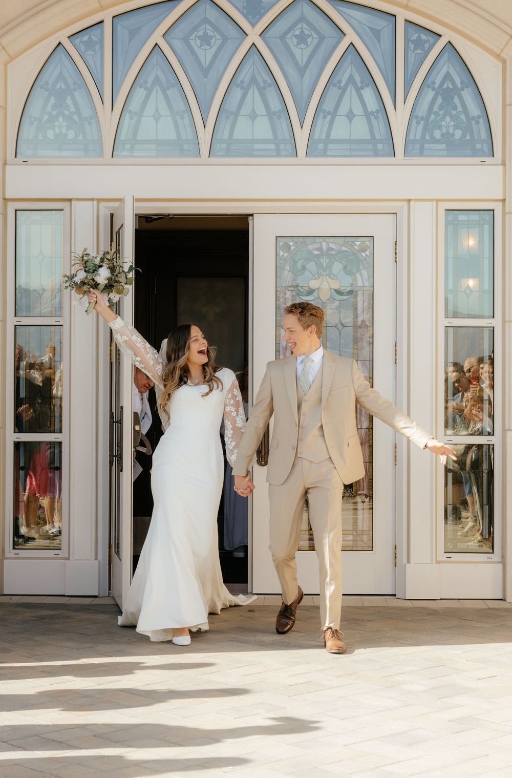 Joyful newlyweds emerging from temple doors, bride raising bouquet in celebration.