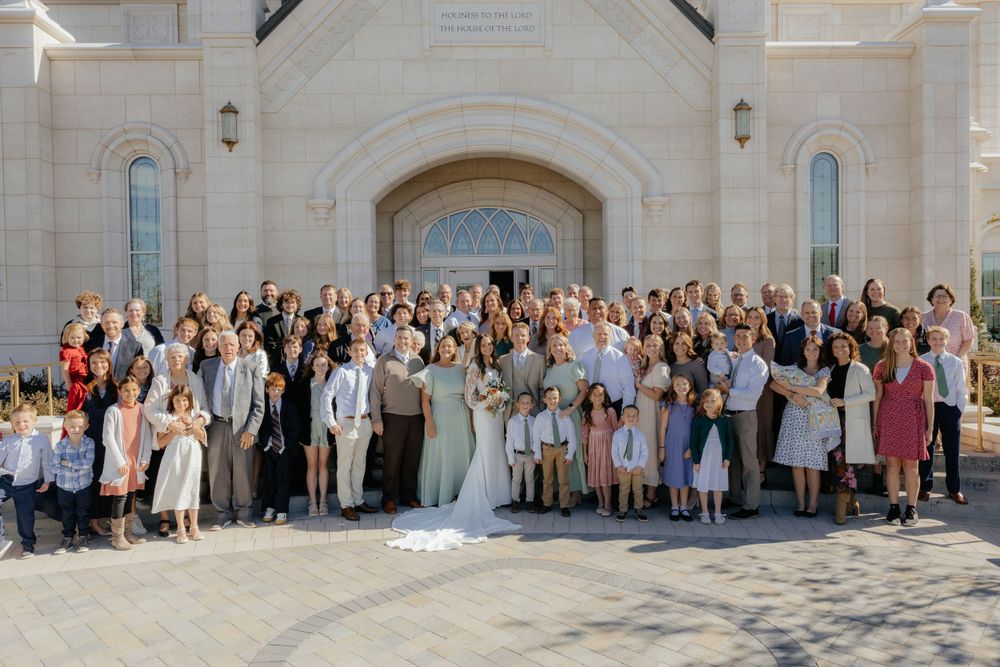 Large wedding party group photo with bride and groom outside an LDS temple on a sunny day.