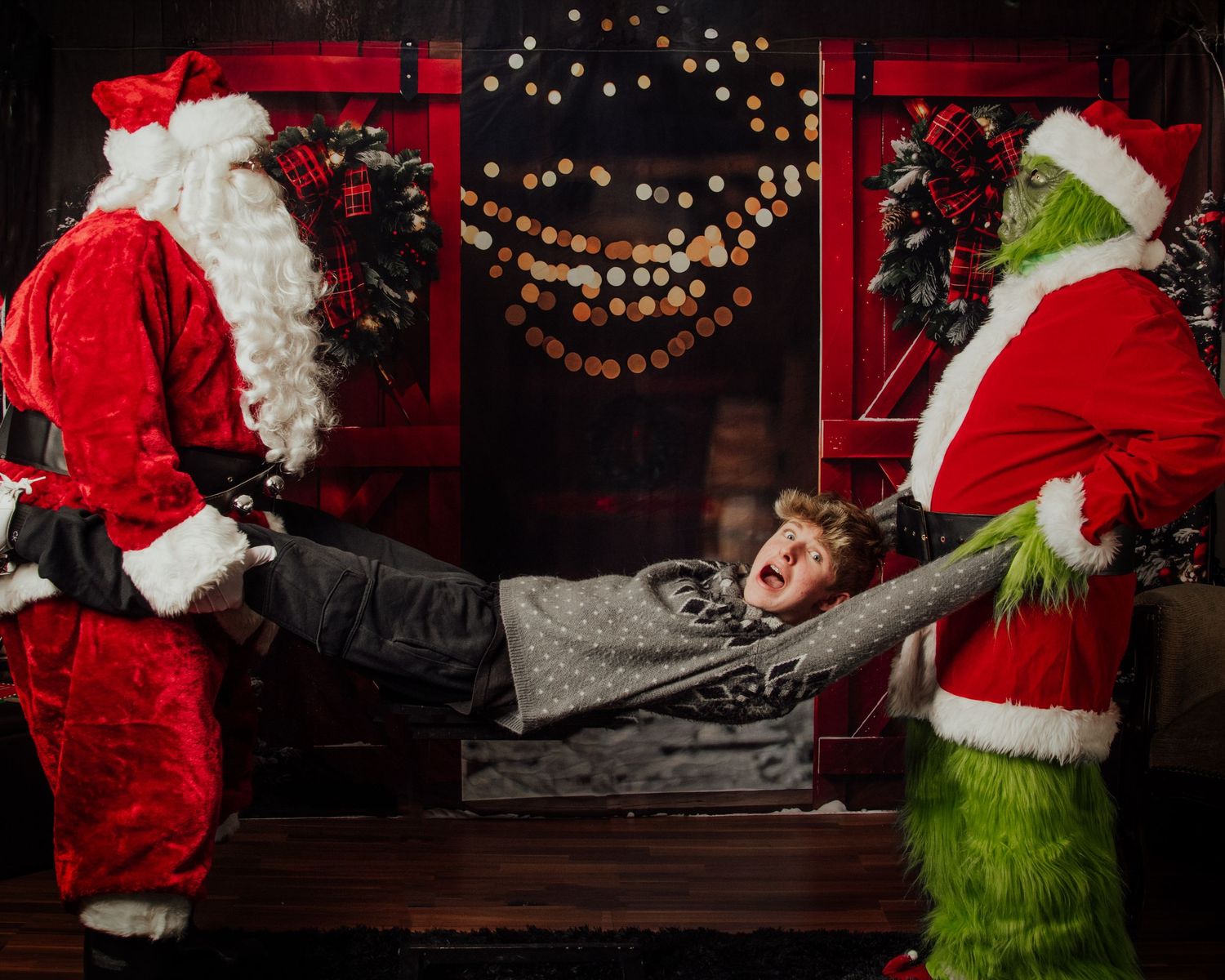 A playful Christmas photo with Santa and the Grinch pulling on opposite ends of a person lying on a bench.