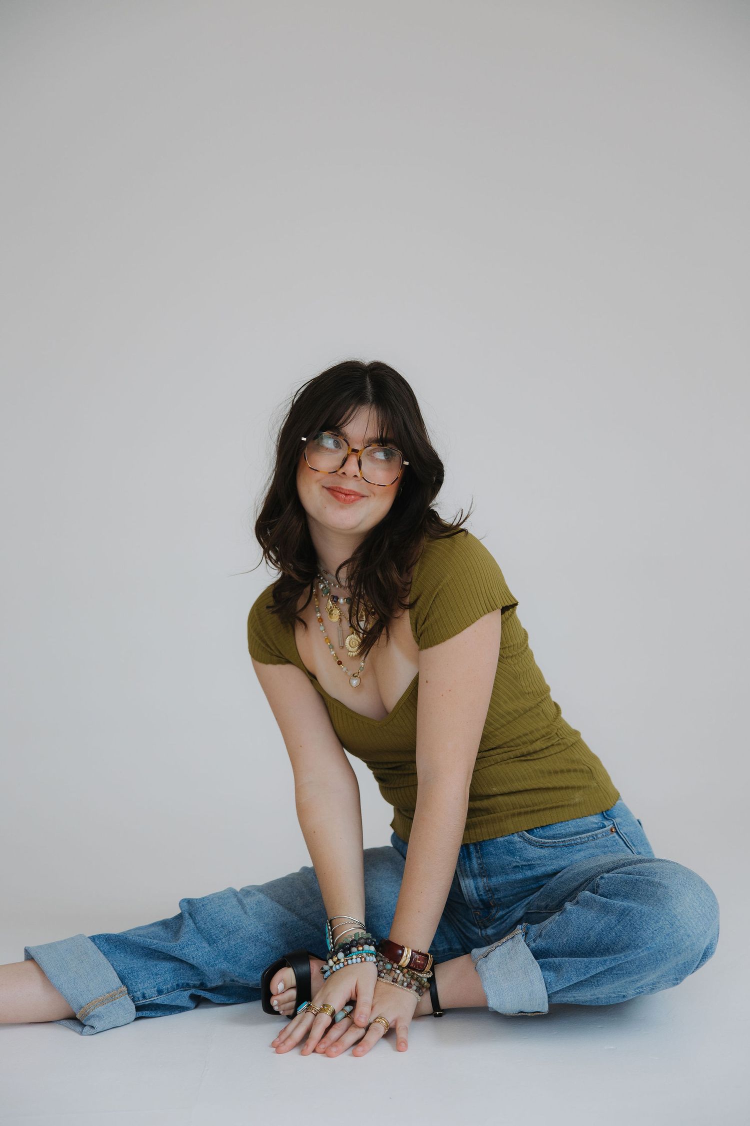 Photographer in olive green top and denim holds professional camera while wearing stylish glasses and jewelry against white wall.