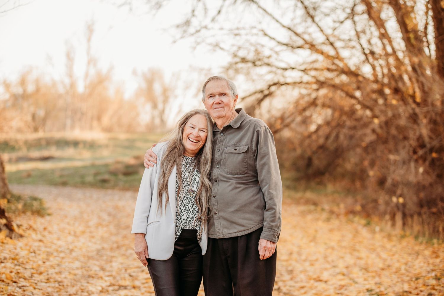 Couple in casual attire embracing while standing among fall foliage.