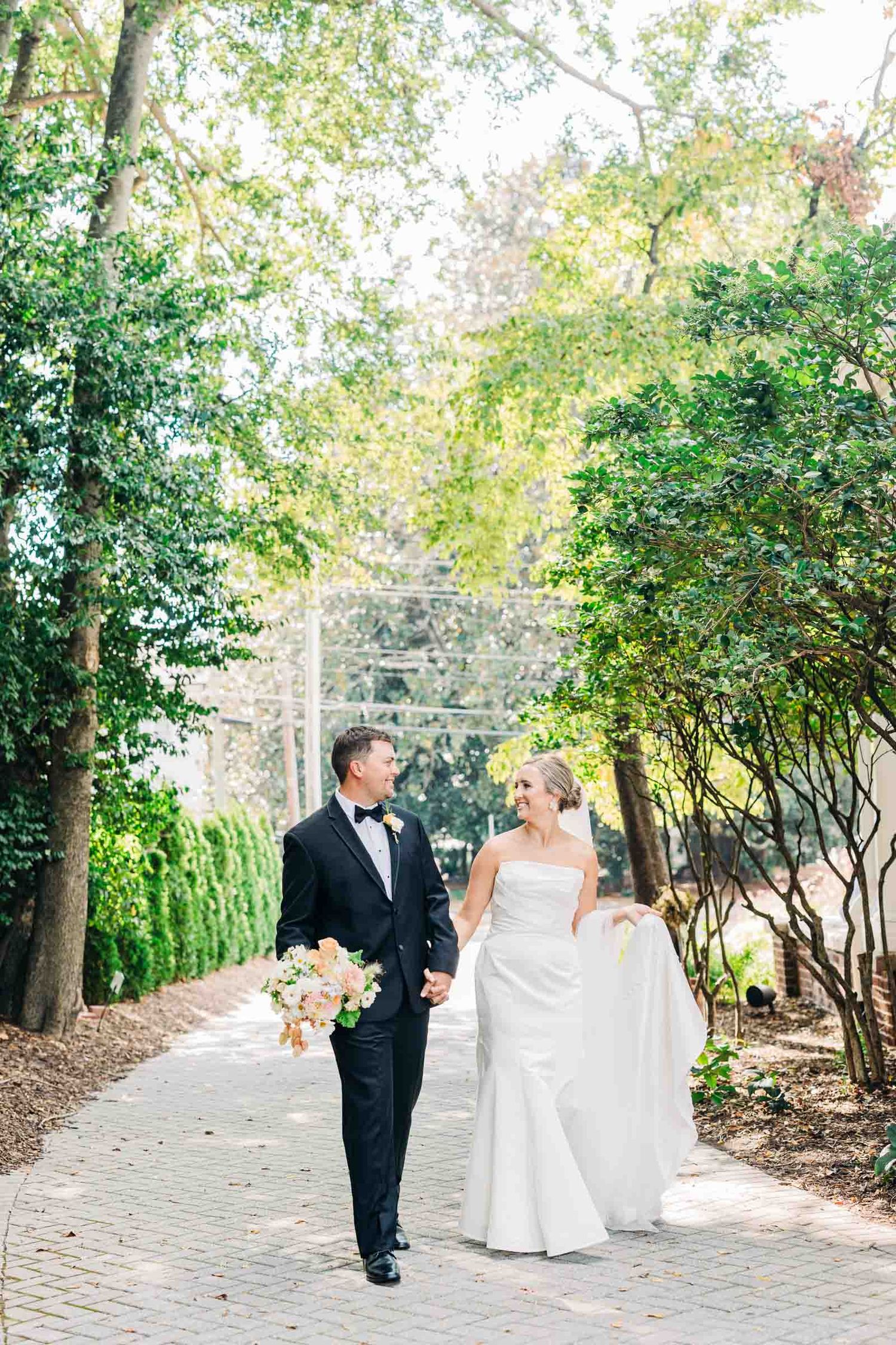 A couple walks together on a brick path surrounded by lush greenery and trees at their outdoor garden wedding 255 Milledge Athens wedding