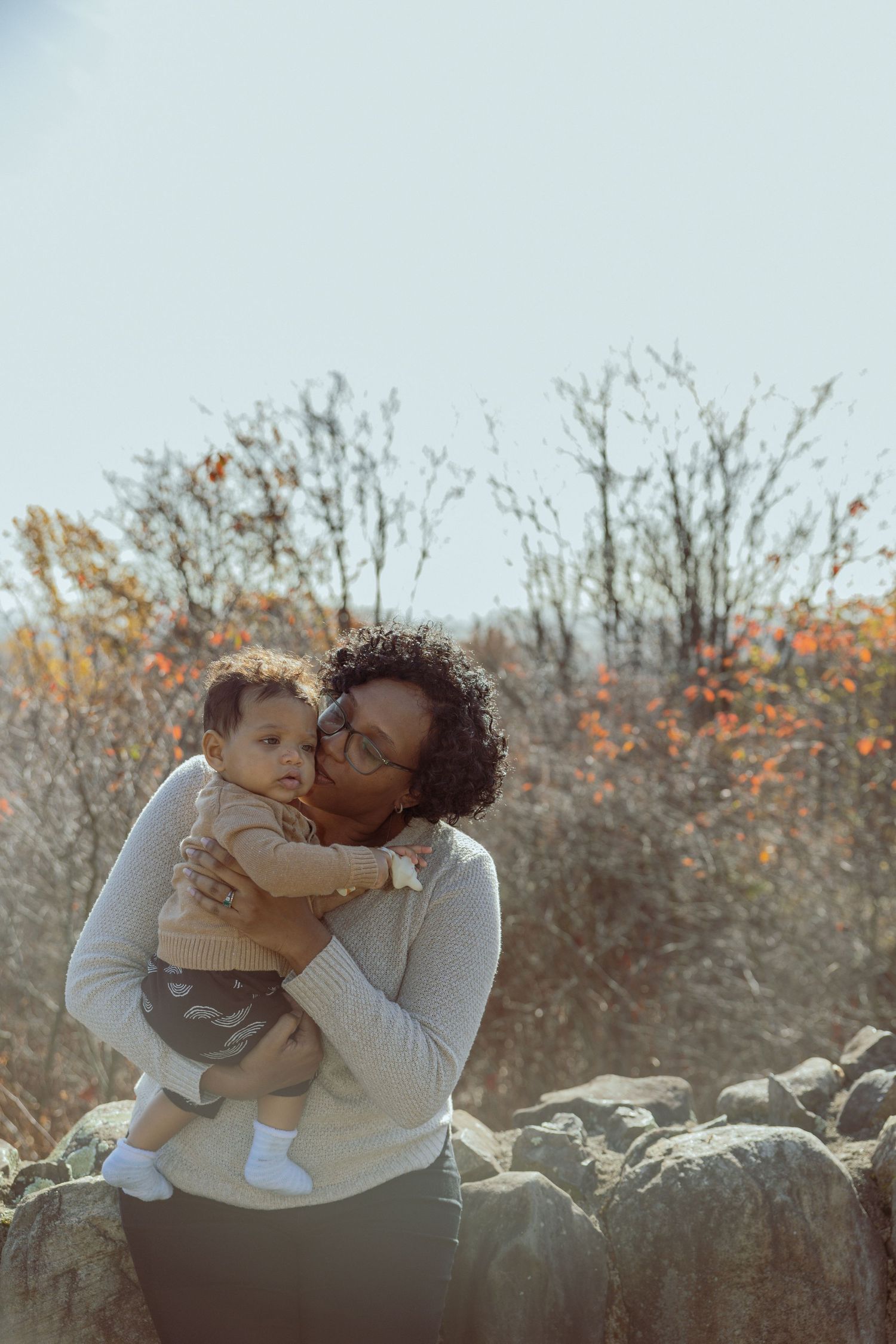 A tender autumn moment captured outdoors as affection is shared while sitting on a rocky ledge with fall foliage in background.