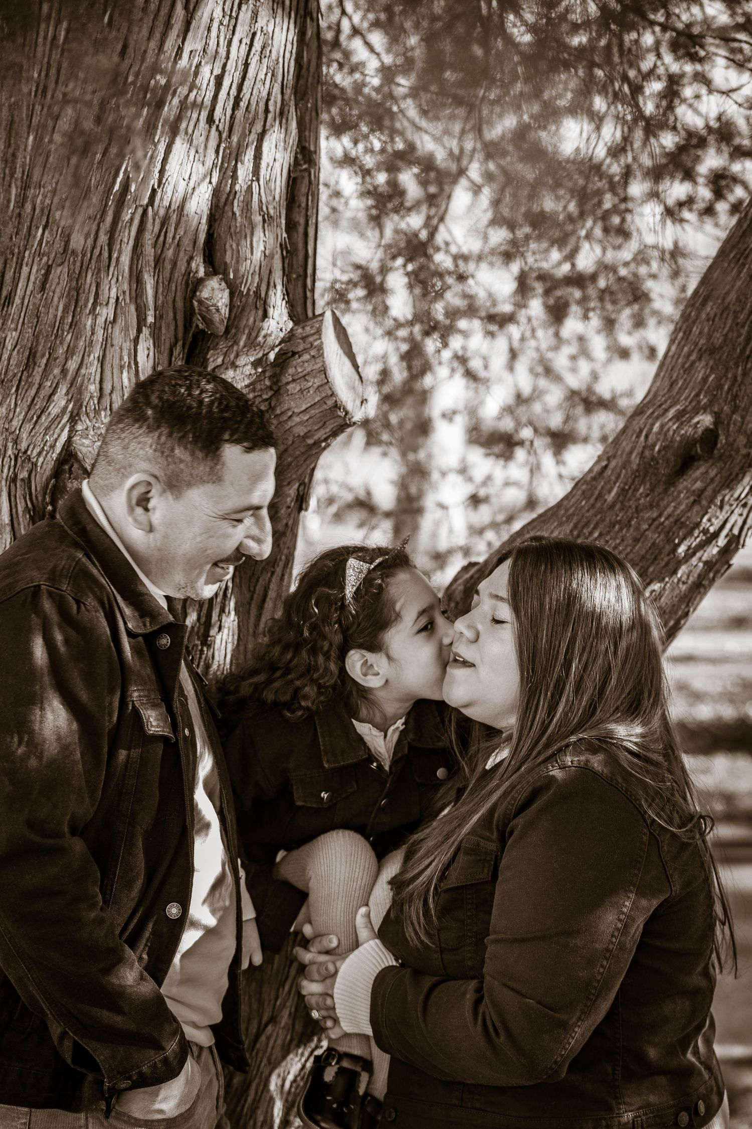 A family of three sharing a tender moment together outdoors in a sepia-toned photograph by a tree.