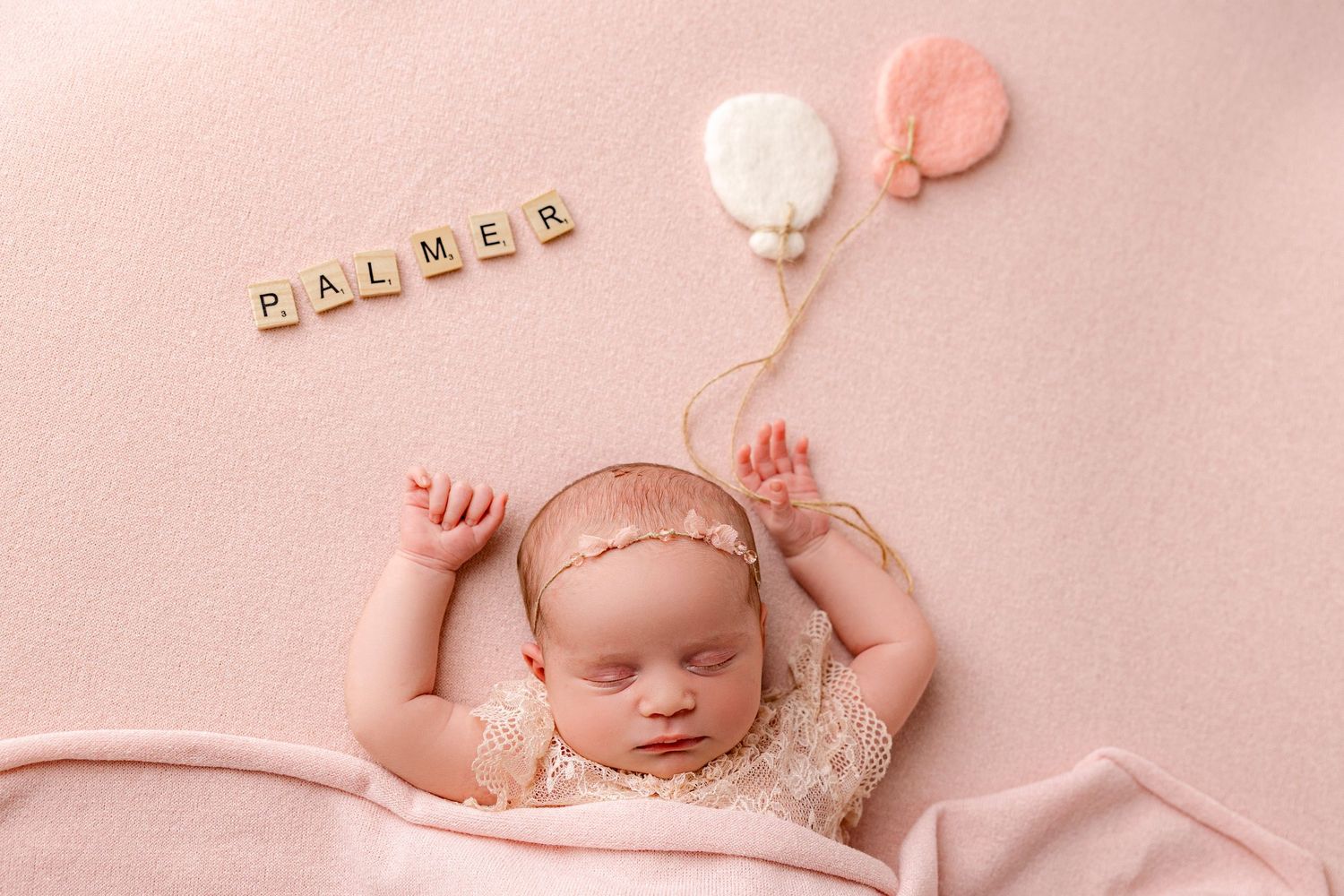A sleeping newborn baby in white lace dress and headband lies peacefully on cream colored fabric.