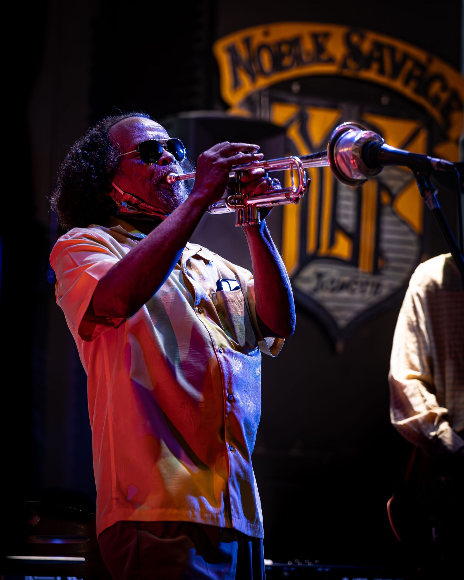 A musician plays trumpet on stage under red lighting at Noble Savage Tavern, silhouetted against the venue's sign in background.