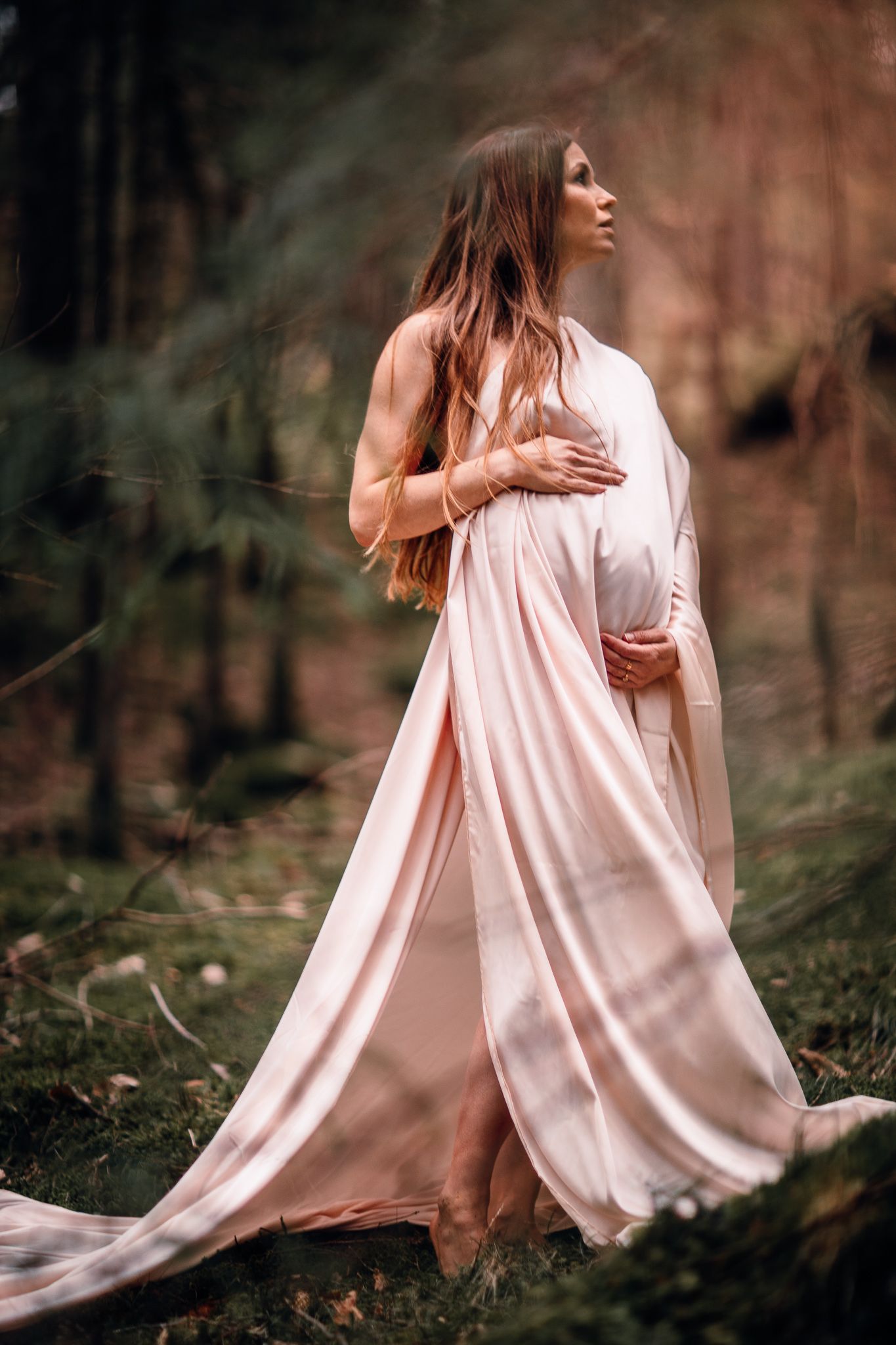 Pregnant woman in flowing pink dress poses in moody forest setting.