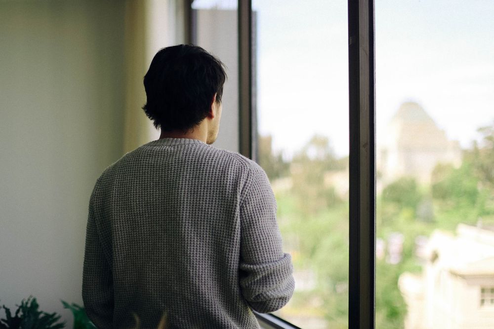 Person in grey sweater looking out large window at urban landscape.