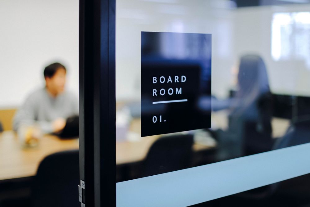 Modern boardroom glass door with black signage and meeting in progress.