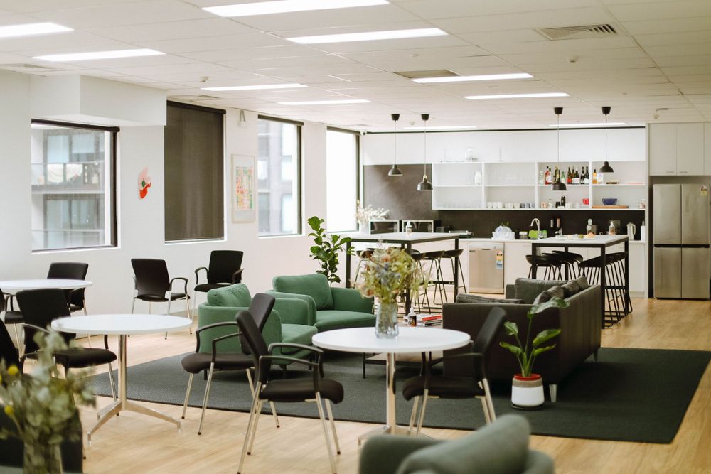 Modern office break room with green chairs and plants near kitchen area.