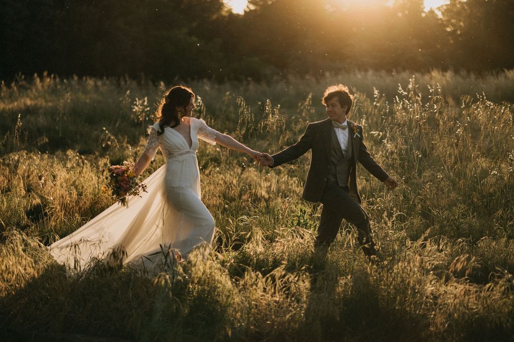 A couple in wedding attire holds hands in a golden sunset-lit meadow with tall grass.