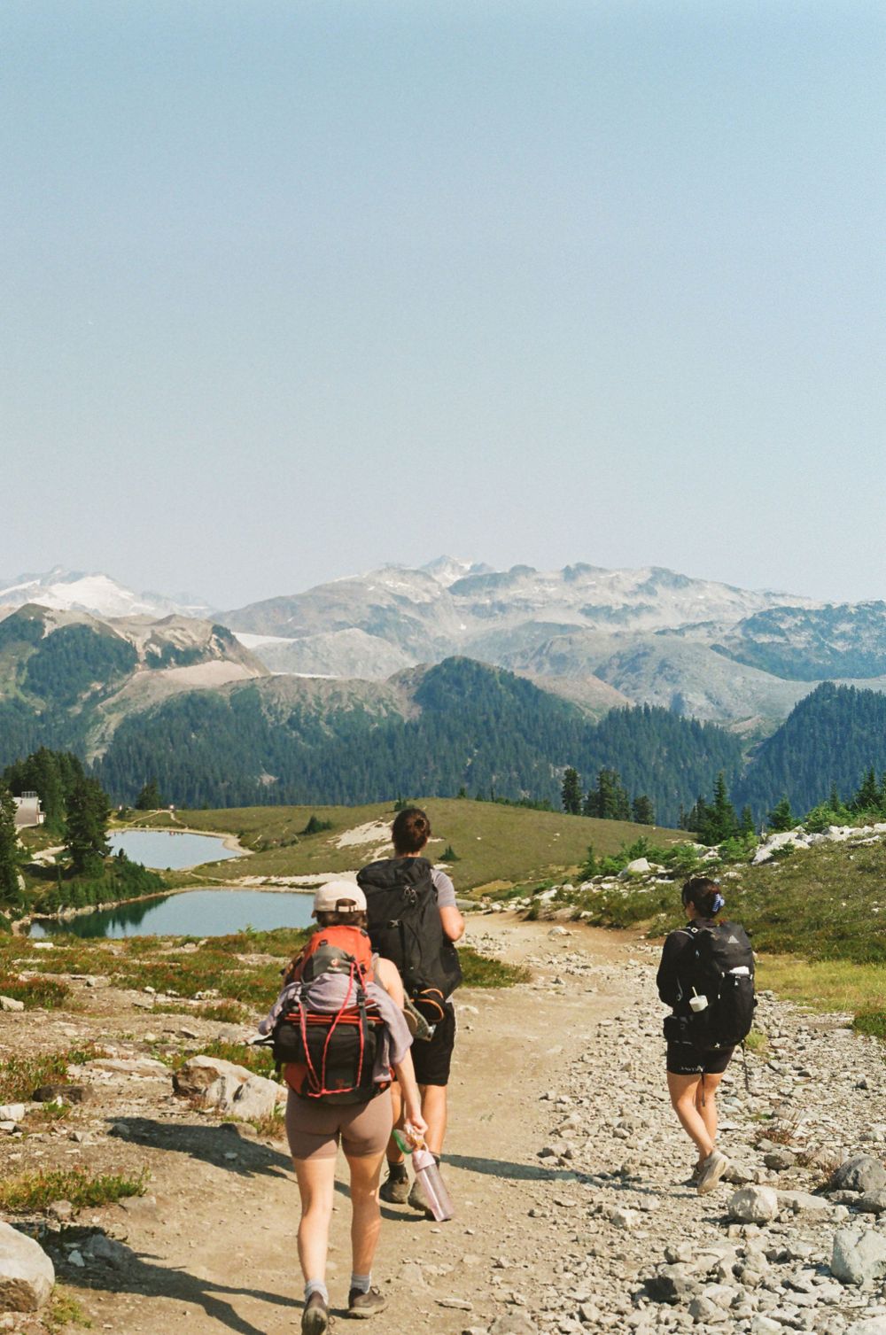 Hikers follow a rocky mountain path with alpine lakes and snow-capped peaks in view.