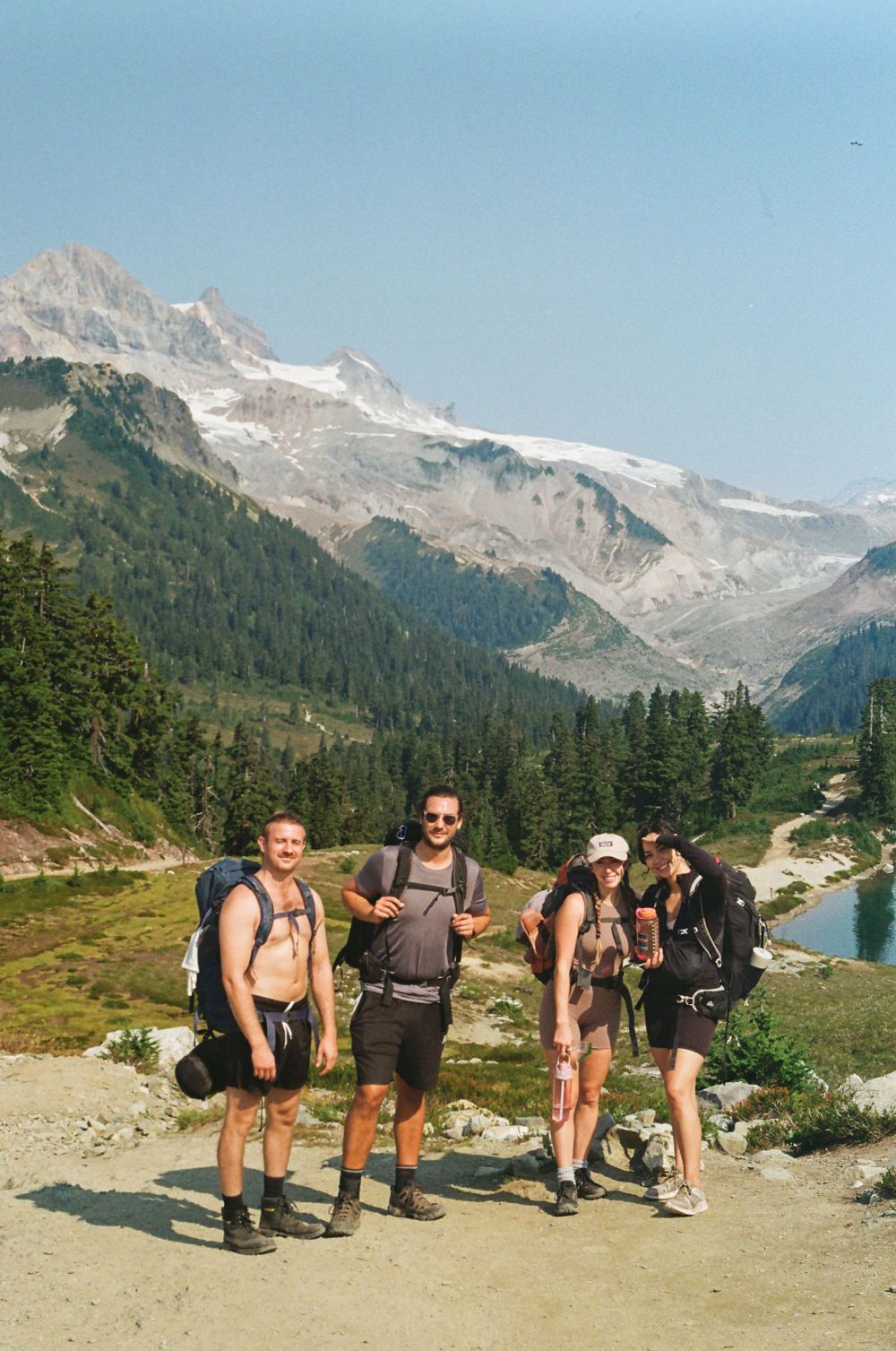 Hikers on a mountain trail with snowy peaks and evergreen forests in the background.