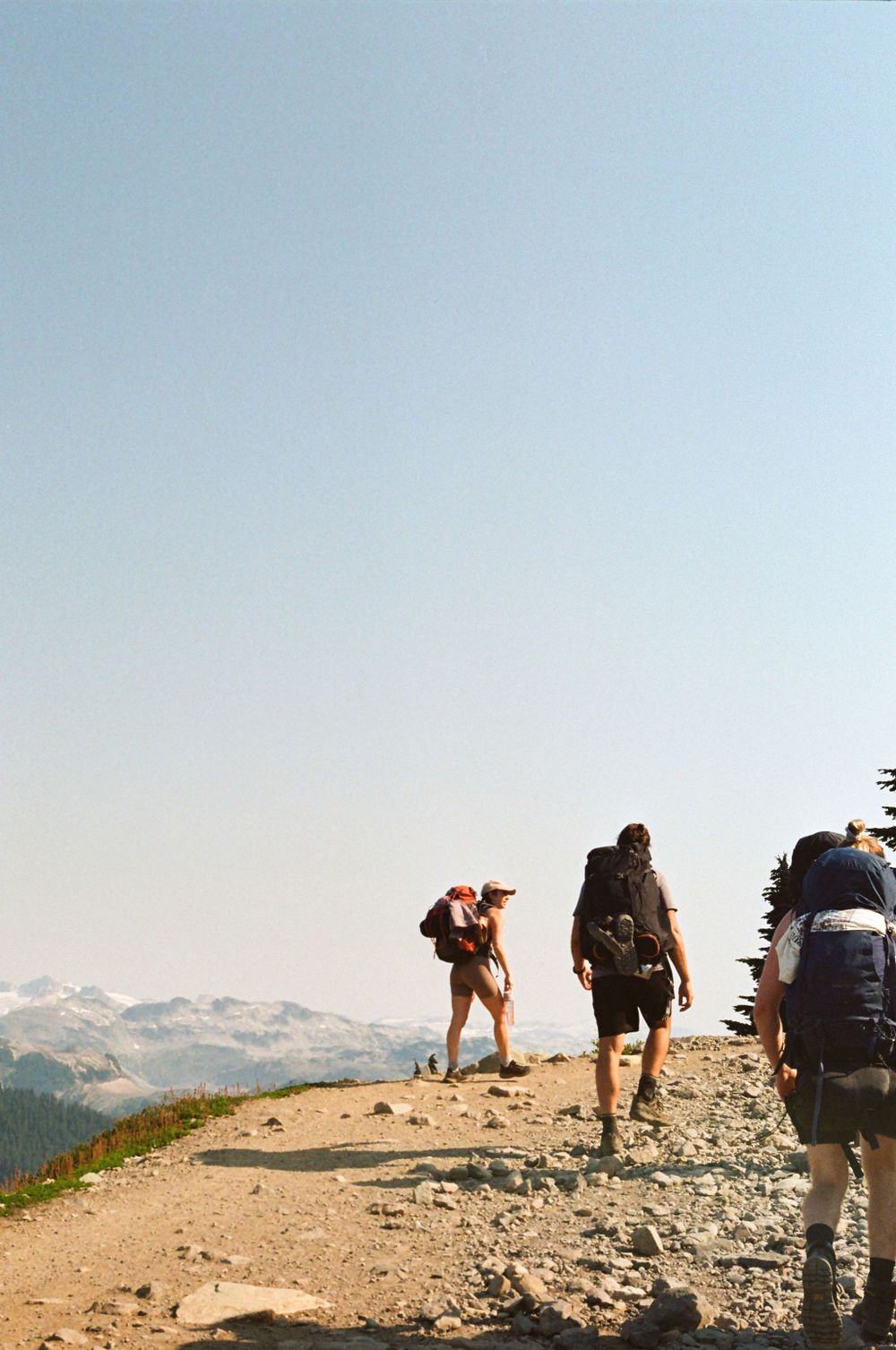 Hikers traverse a rocky mountain trail with snow-capped peaks in the distance.