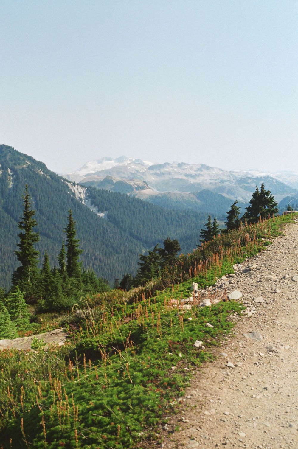A winding mountain trail cuts through alpine terrain with snow-capped peaks visible in the distance.