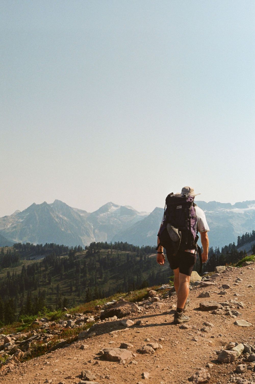 Scenic mountain hiking trail with distant snow-covered peaks and dense forest below.