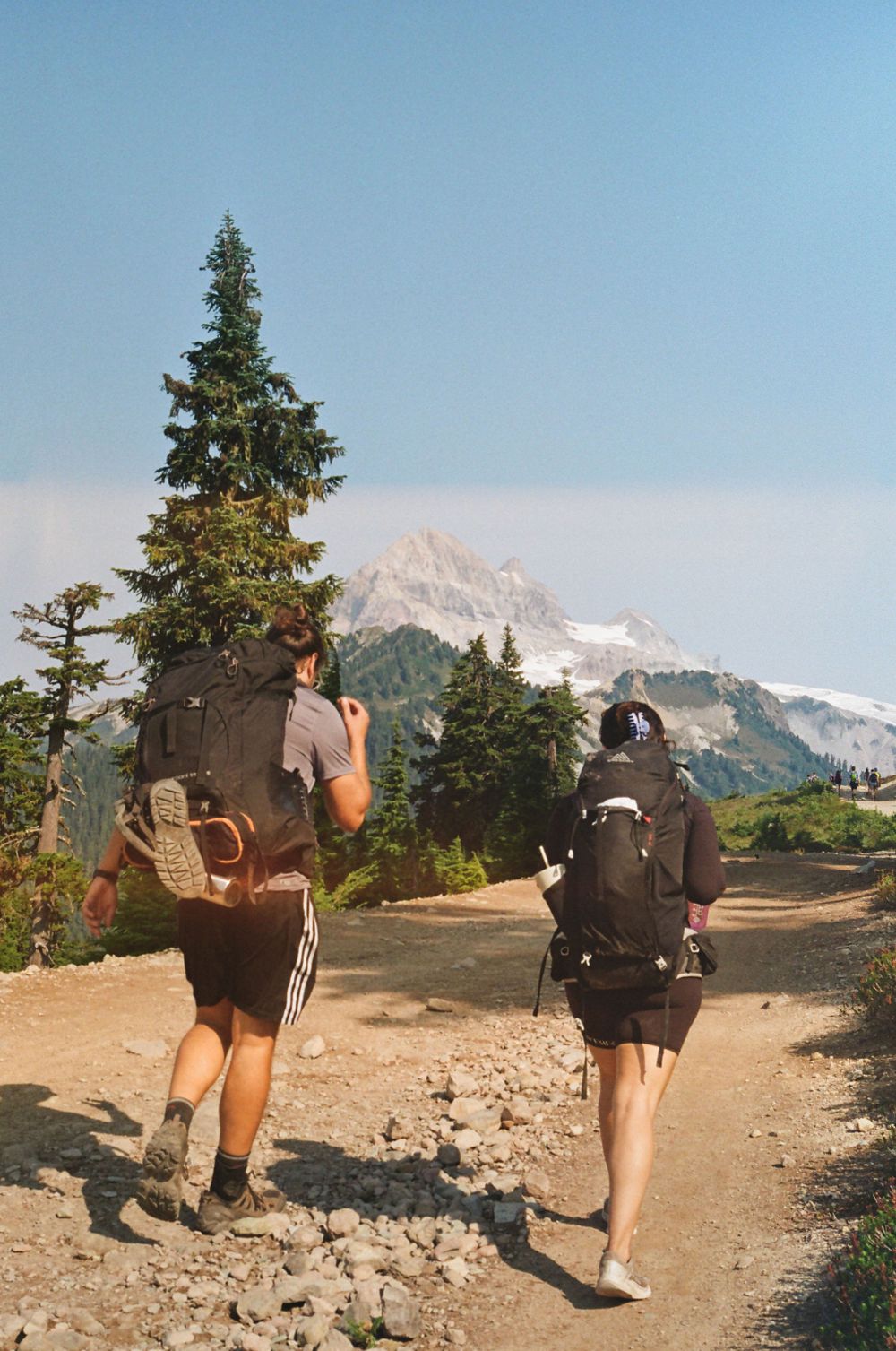 Group of backpackers hiking along a dusty mountain trail with snow-capped peaks in view.