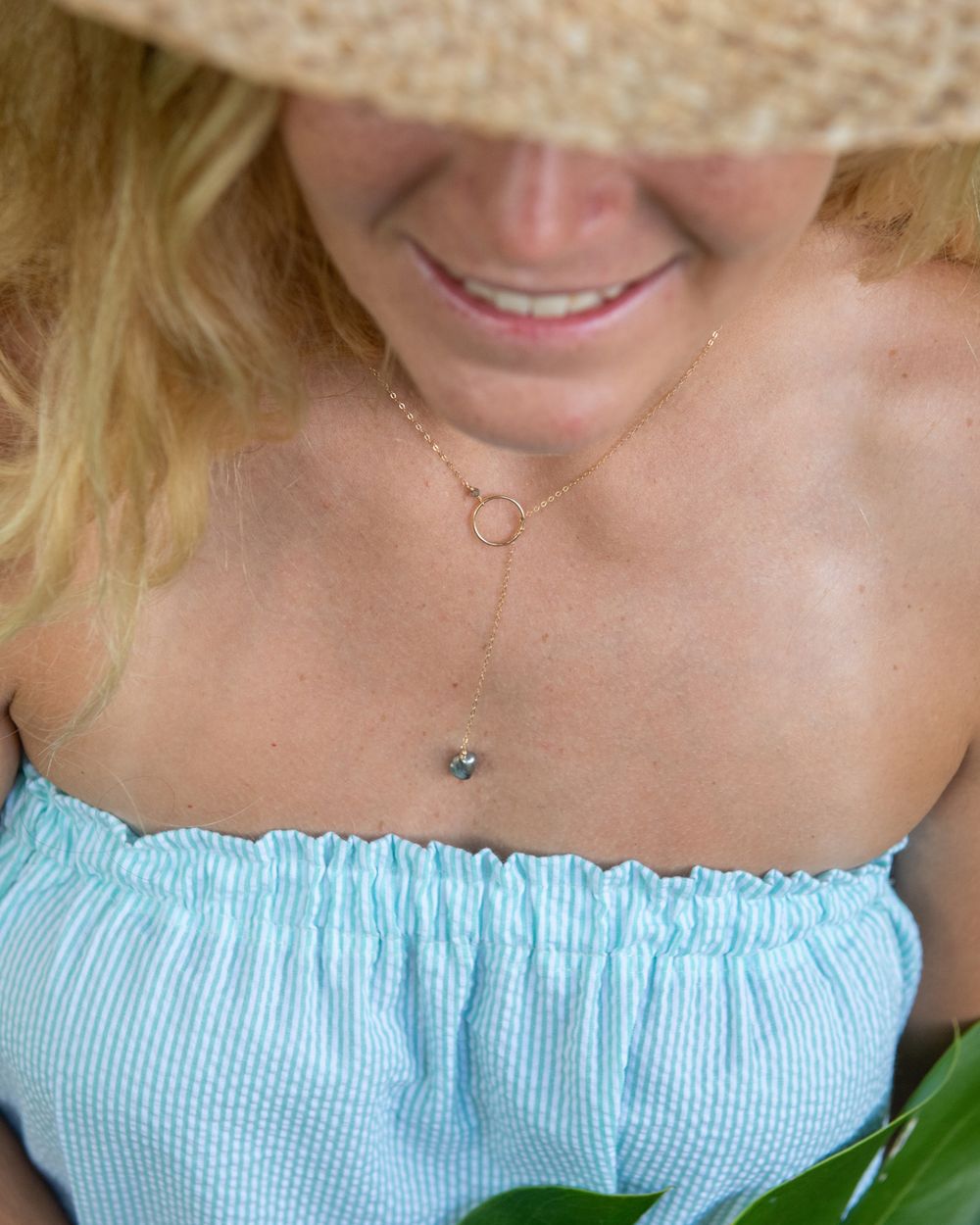 Delicate gold necklace with circular pendant shown on a light blue summer top with straw hat detail.