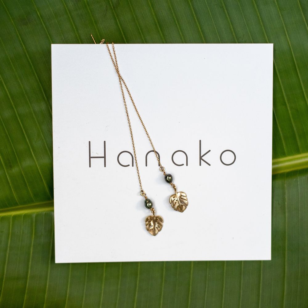 Hanako branded jewelry display with delicate pendant earrings on a tropical leaf background.