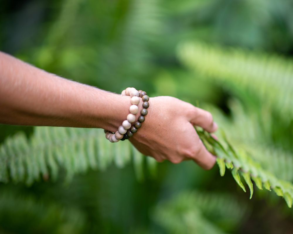 Hand wearing pearl bracelet reaching out to touch a fern leaf in garden.