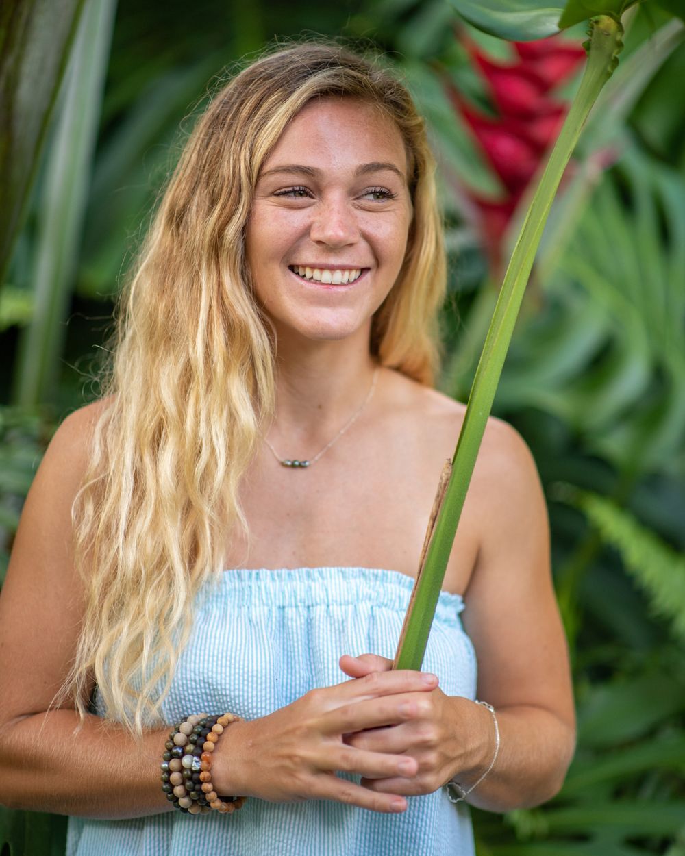 Woman in light blue top posing with tropical plants and flowers.