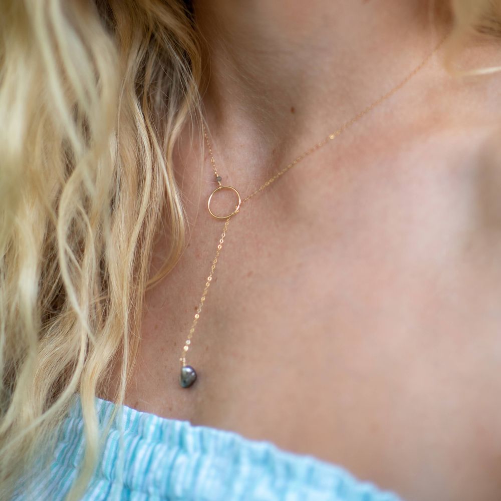Dainty gold necklace with pendant and crystal drop worn against light blue top.