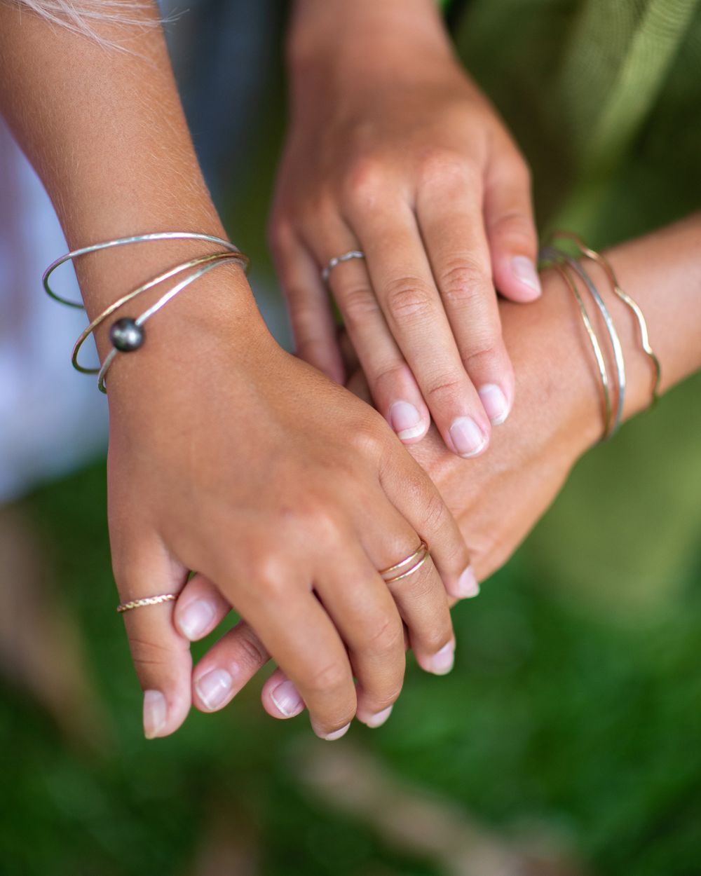 Hands adorned with silver bracelets and rings crossed over green foliage.