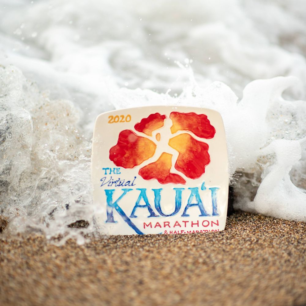 Kauai Marathon logo with red hibiscus design placed on beach sand with gentle surf in background.