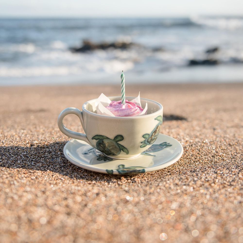 Decorative teacup with sea turtle design and whipped cream sitting on beach sand with ocean view.