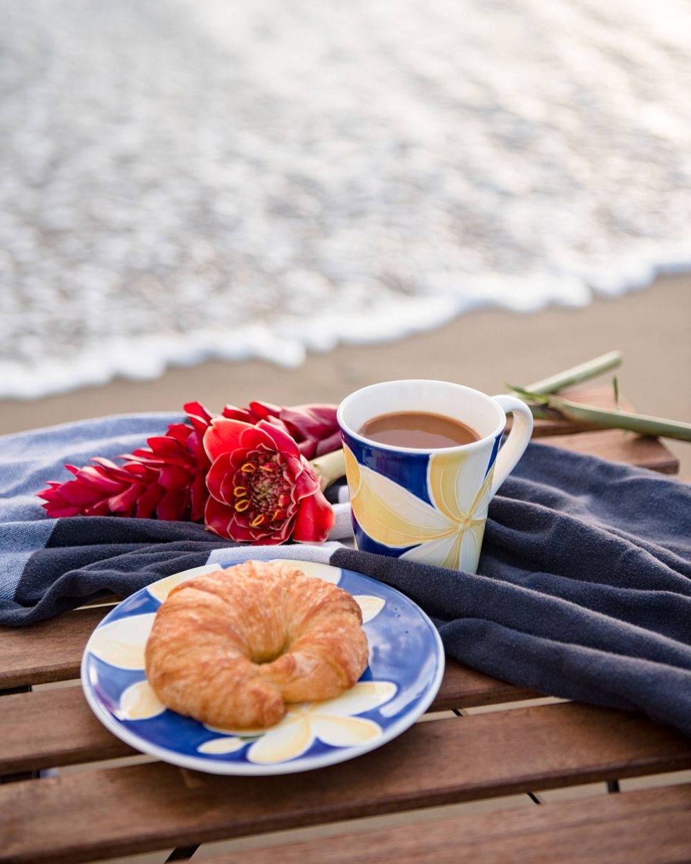 Breakfast scene on beach with croissant, coffee, and red tropical flowers on blue plates against ocean backdrop.