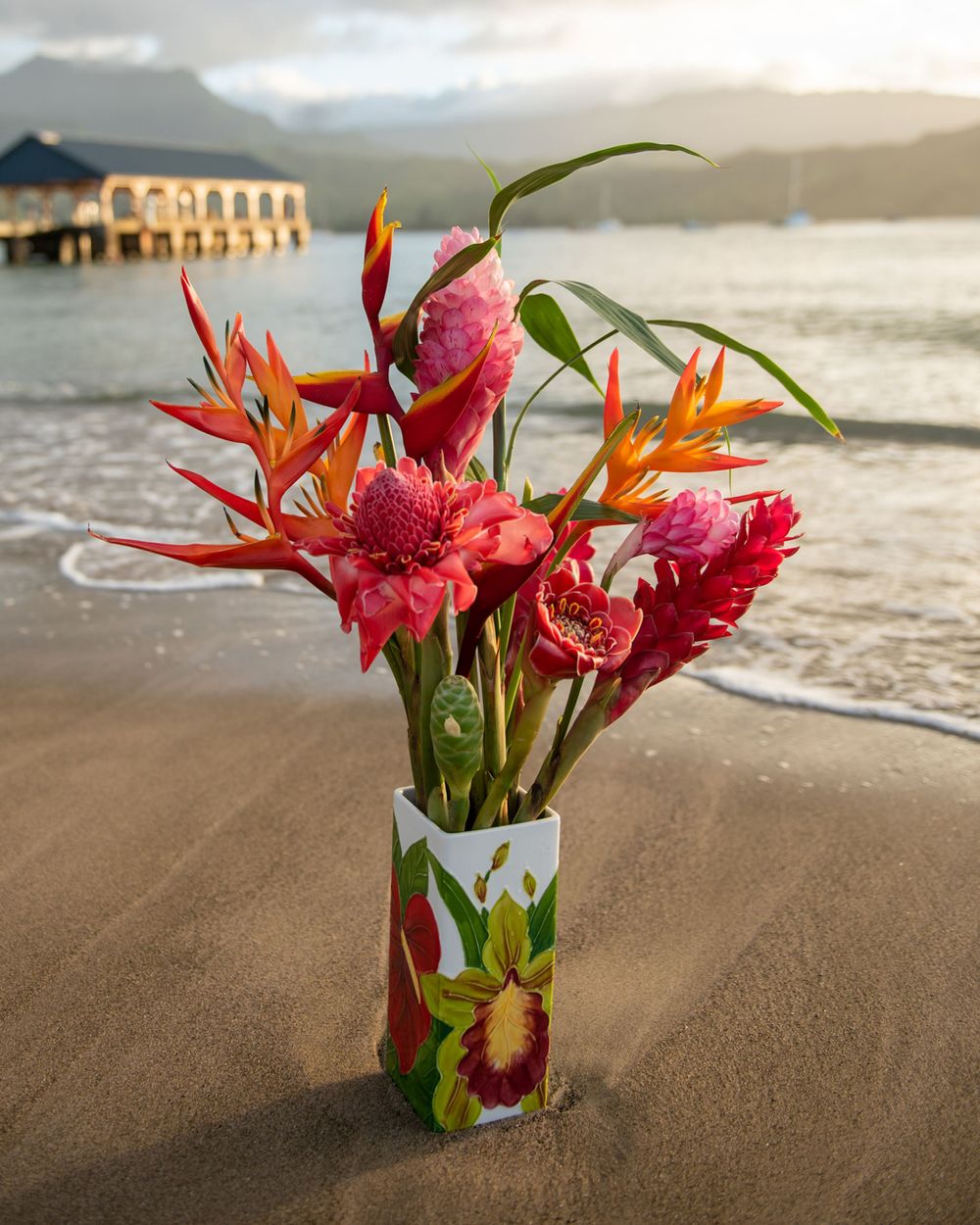 Tropical flower arrangement with birds of paradise and ginger in vase on beach with pier in background.