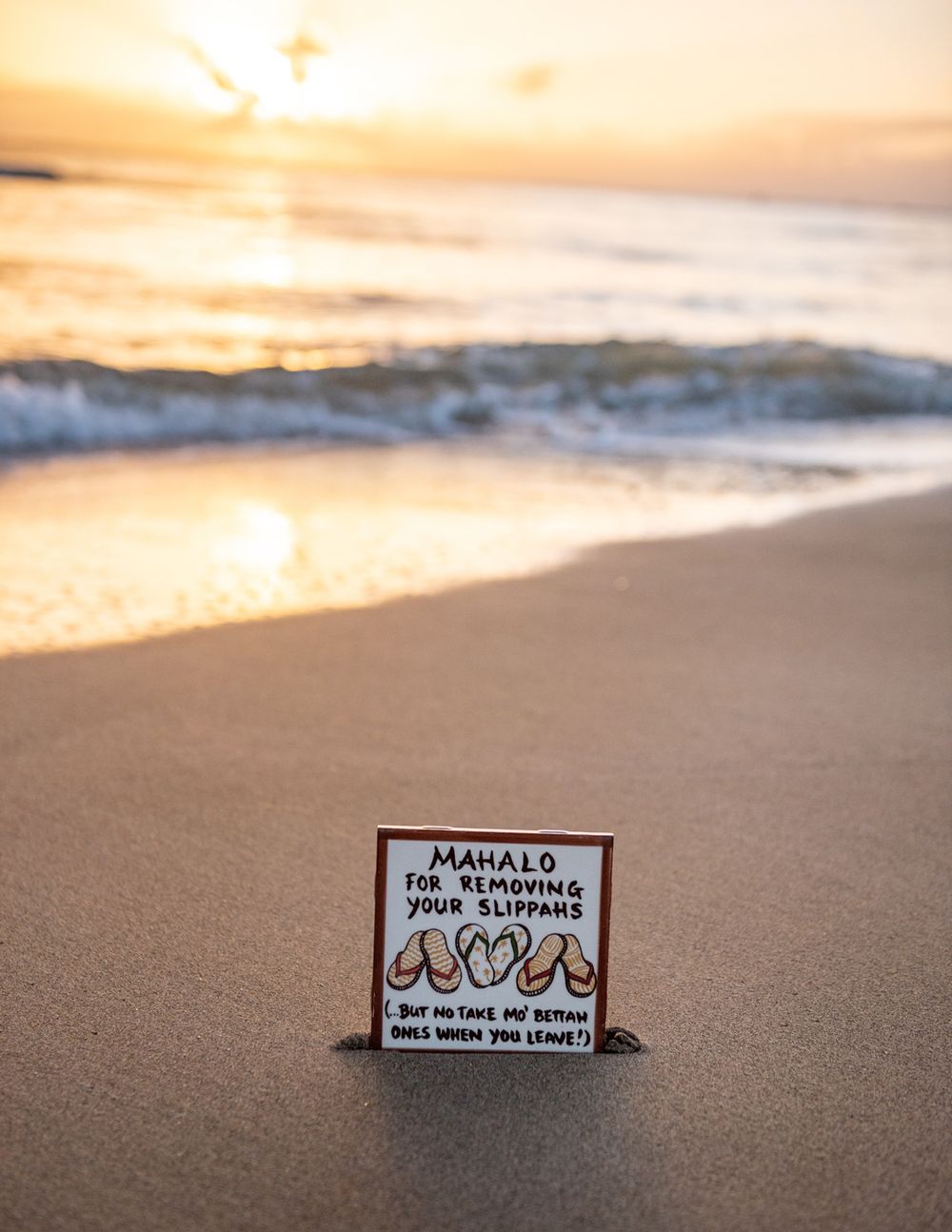 Mahalo thank you sign photographed on sandy beach at sunset with waves in background.