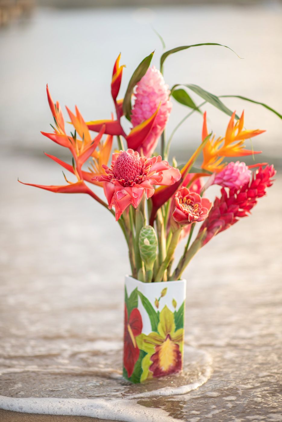 Colorful tropical flower arrangement featuring birds of paradise and ginger in decorative vase on wet sand.