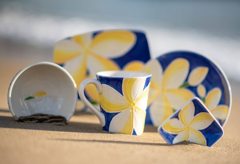 Blue and yellow plumeria print dishes and cups arranged artistically on beach sand.
