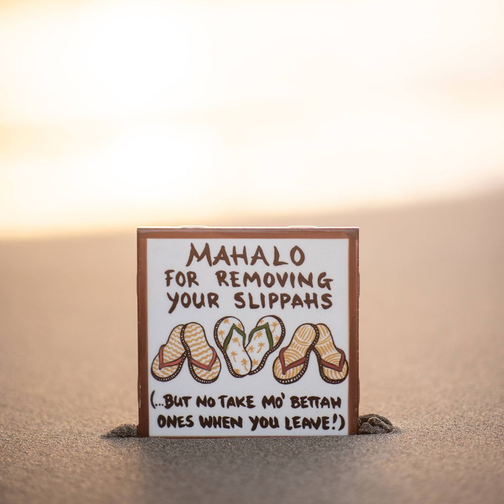 A wooden sign in sand reads 'Mahalo for removing your slippings' with flip flop illustrations.