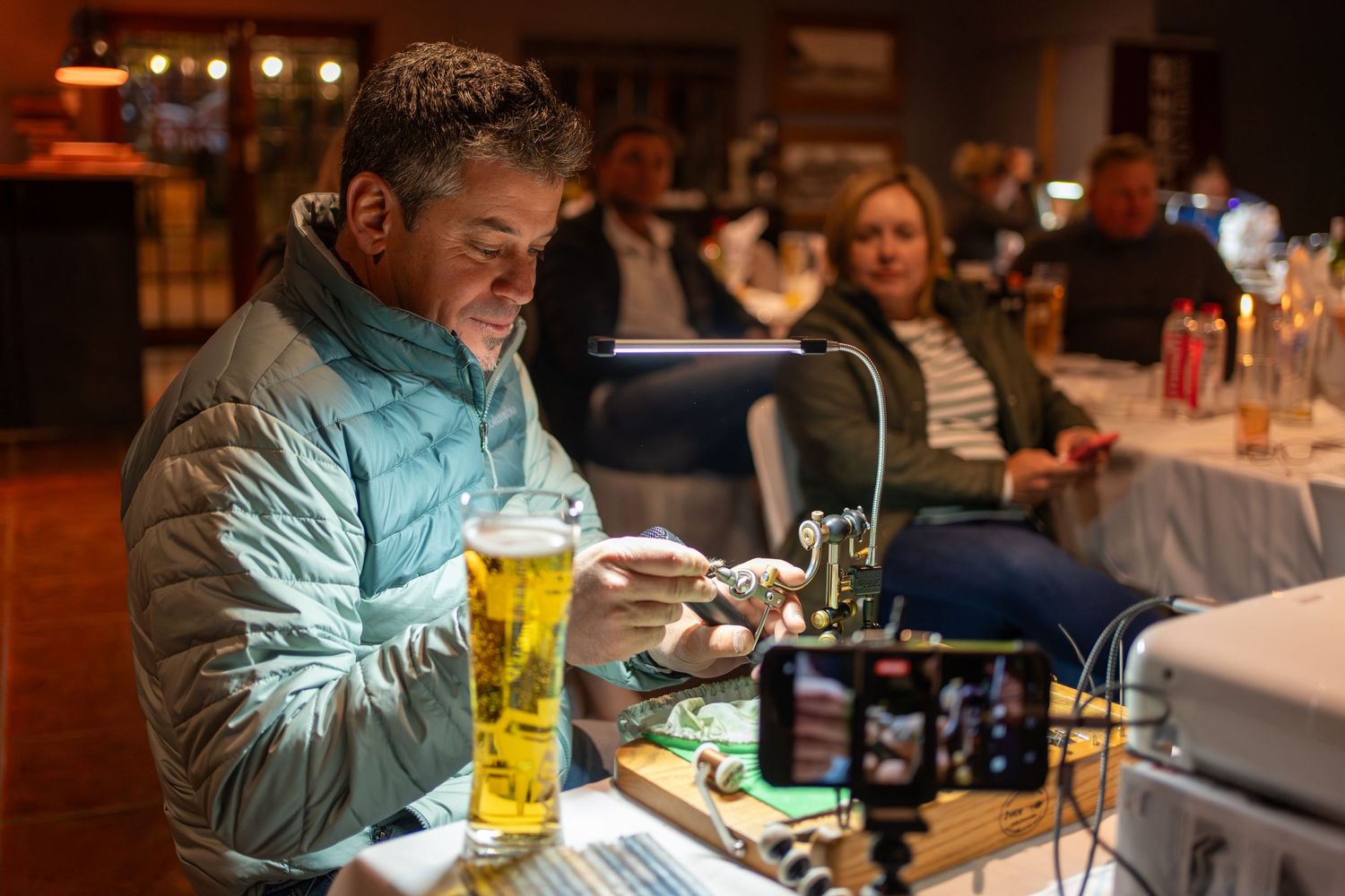 Person in blue jacket looking at phone with beer glass at table in dimly lit room.
