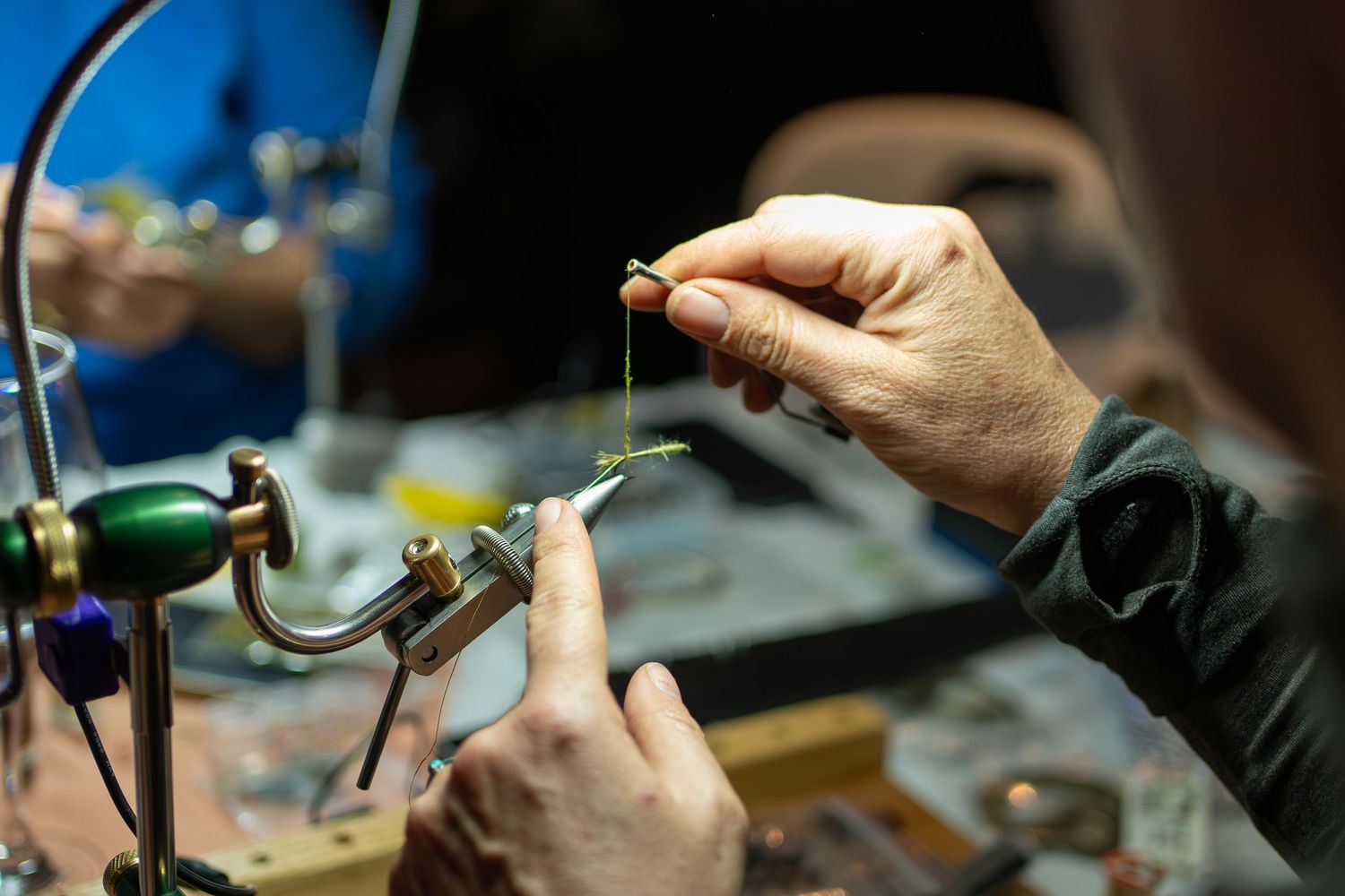 Close-up of hands tying a fishing fly at a workbench with tools.