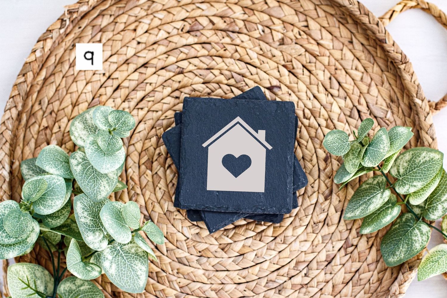 Simple house icon slate coaster displayed on woven rattan mat with green leaf decoration.