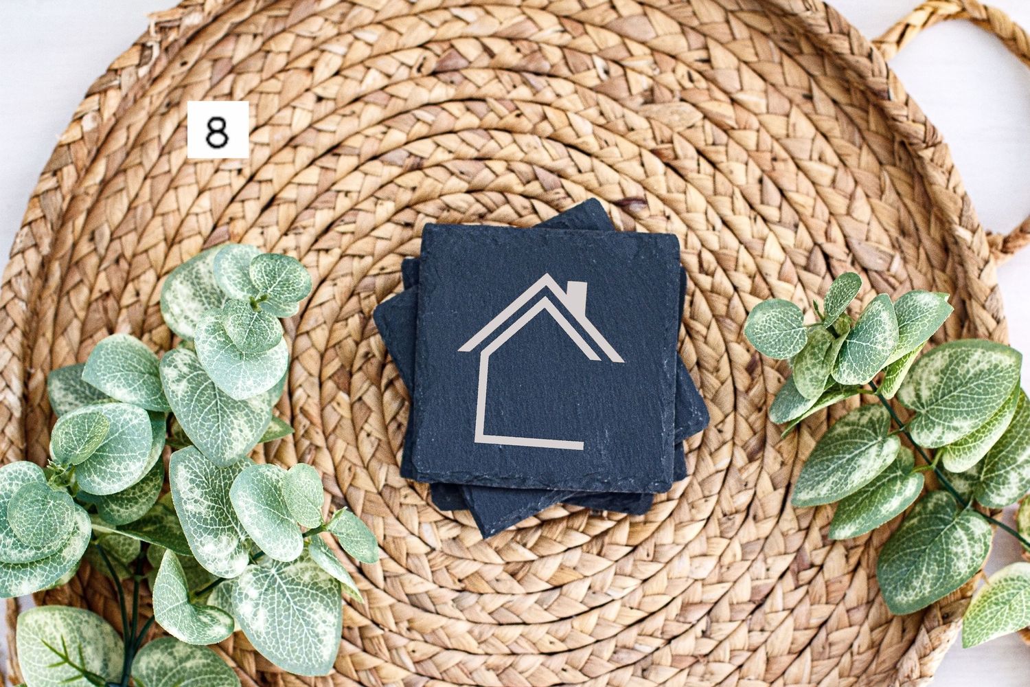 Navy blue house icon tag displayed on circular woven jute mat with sage green artificial eucalyptus leaves.