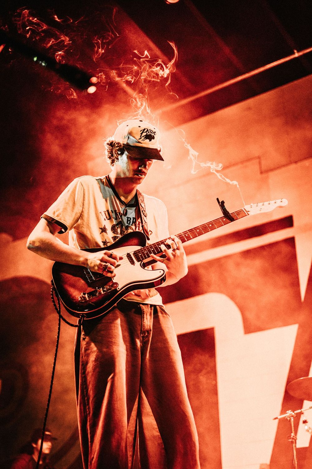 A musician performs on stage with an electric guitar amid dramatic red lighting and smoke effects.